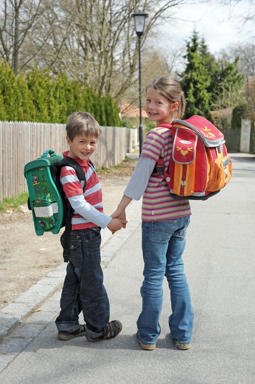 Ein Schuljunge und ein Schulmädchen mit Ränzen auf den Rücken halten sich an den Händen, in die Kamera blickend, auf dem Weg zur Schule