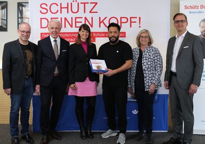 die Projektbeteiligten stehen vor dem Banner Schütz Deinen Kopf - Gehirnerschütterungen in der Schule vermeiden, mittig mit der Box steht Adel Tawil als Präsident der ZNS