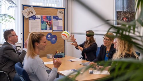 Projektverantwortliche Sam Westphal und Leonie Schulte sitzen mit dem stv. Geschäftsführer des GUV OL, Henning Wolff, Theresa Wieting, Präventionsberaterin und Johanna Verse, Öffentlichkeitsarbeit beim GUV OL den Einsatz von freiwilligen Helfenden beim Host Town 2023. Henning Wolff wirft Theresa Wieting, die eine Sichtschutzbrille trägt, einen Ball zu