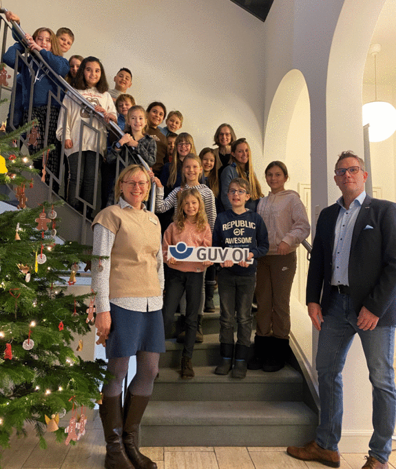 Viertklässlerinnen und Viertklässler stehen auf der Treppe mit dem GUV OL - Schild in der Hand zusammen mit ihrer Lehrerin Christine Krüger und Angela Pauka und Henning Wolff vom GUV OL 