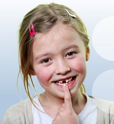 Mädchen im Grundschulalter hält einen Finger auf ihren fehlenden Frontschneidezahn, lächelt
