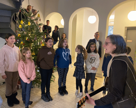 Kinder stehen singend vorm Weihnachtsbaum im Foyer. Ihre Lehrerin spielt dazu Gitarre. 