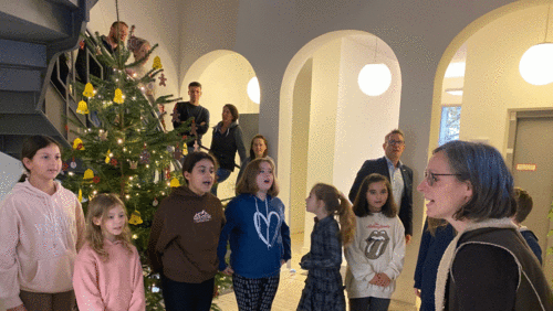 Kinder stehen singend vorm Weihnachtsbaum im Foyer. Ihre Lehrerin spielt dazu Gitarre. 