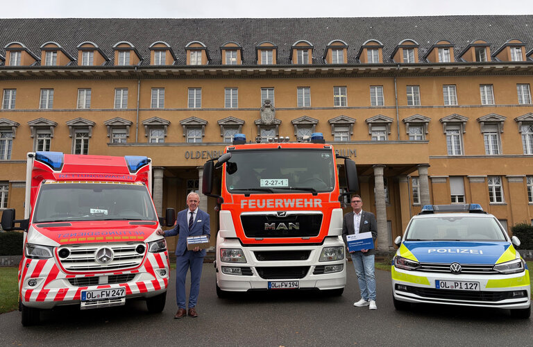 Michael May, Geschäftsführer des GUV OL, und Henning Wolff, stv. Geschäftsführer, stellen die Karte für Ersthelfende vor dem Alten Oldenburgischen Landtag vor, flankiert von Fahrzeugen des Rettungsdienstes, der Polizei und der Feuerwehr Oldenburg