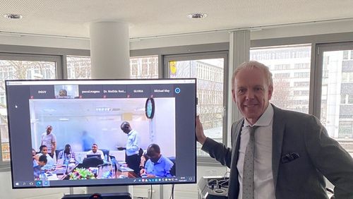 Michael May, Geschäftsführer des GUV OL, im Berliner Besprechungsraum der VBG, vor einem Monitor stehend, der die Teilnehmenden des Trainings aus Tansania zeigt