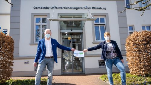 Michael May und Johanna Verse, beide in Jeans und Blazer, zeigen das Jubiläumslogo 50 Jahre SUV vor dem Dienstgebäude des GUV OL 