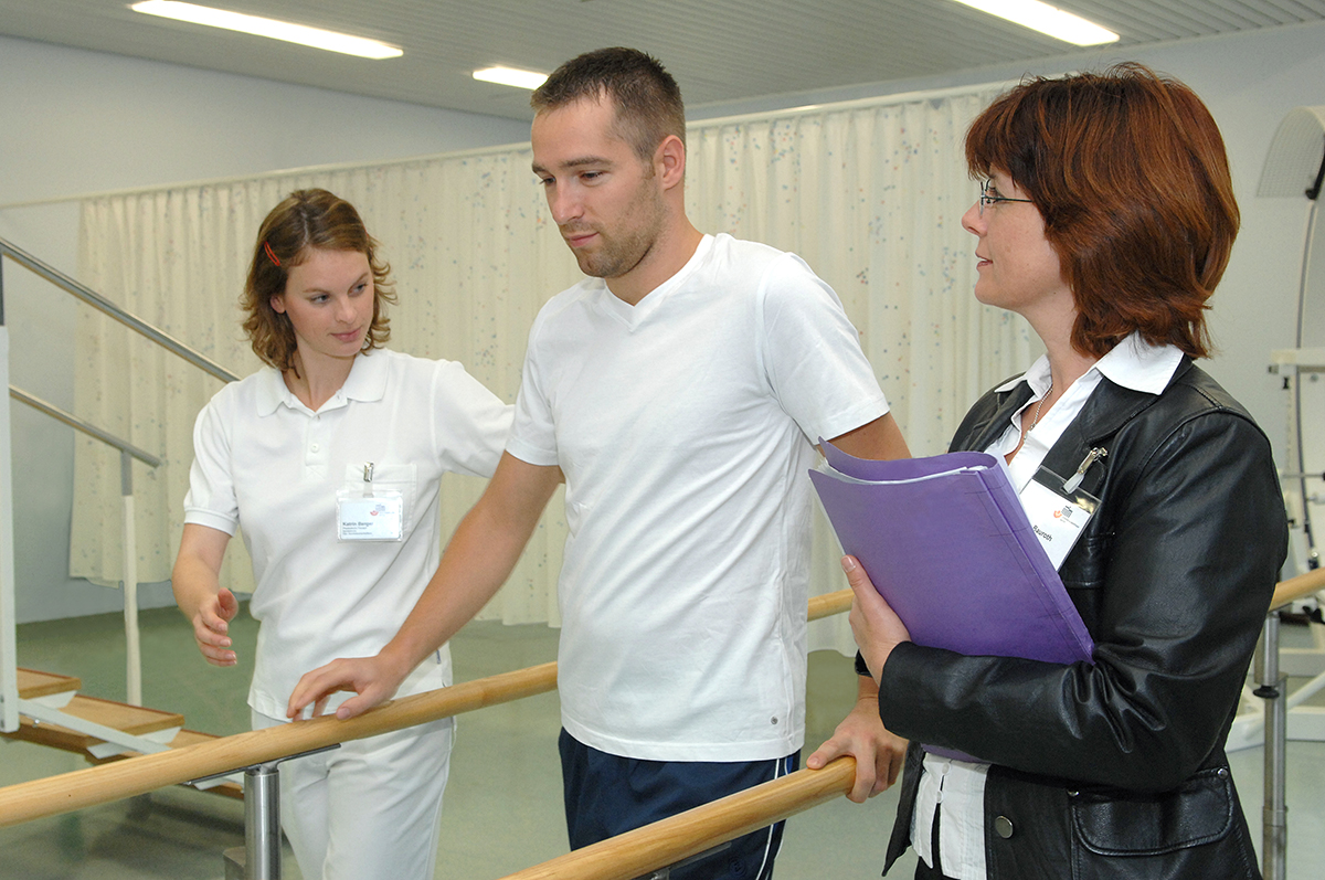 Ein Reha-Patient steht zwischen zwei Mitarbeiterinnen einer Reha-Einrichtung 