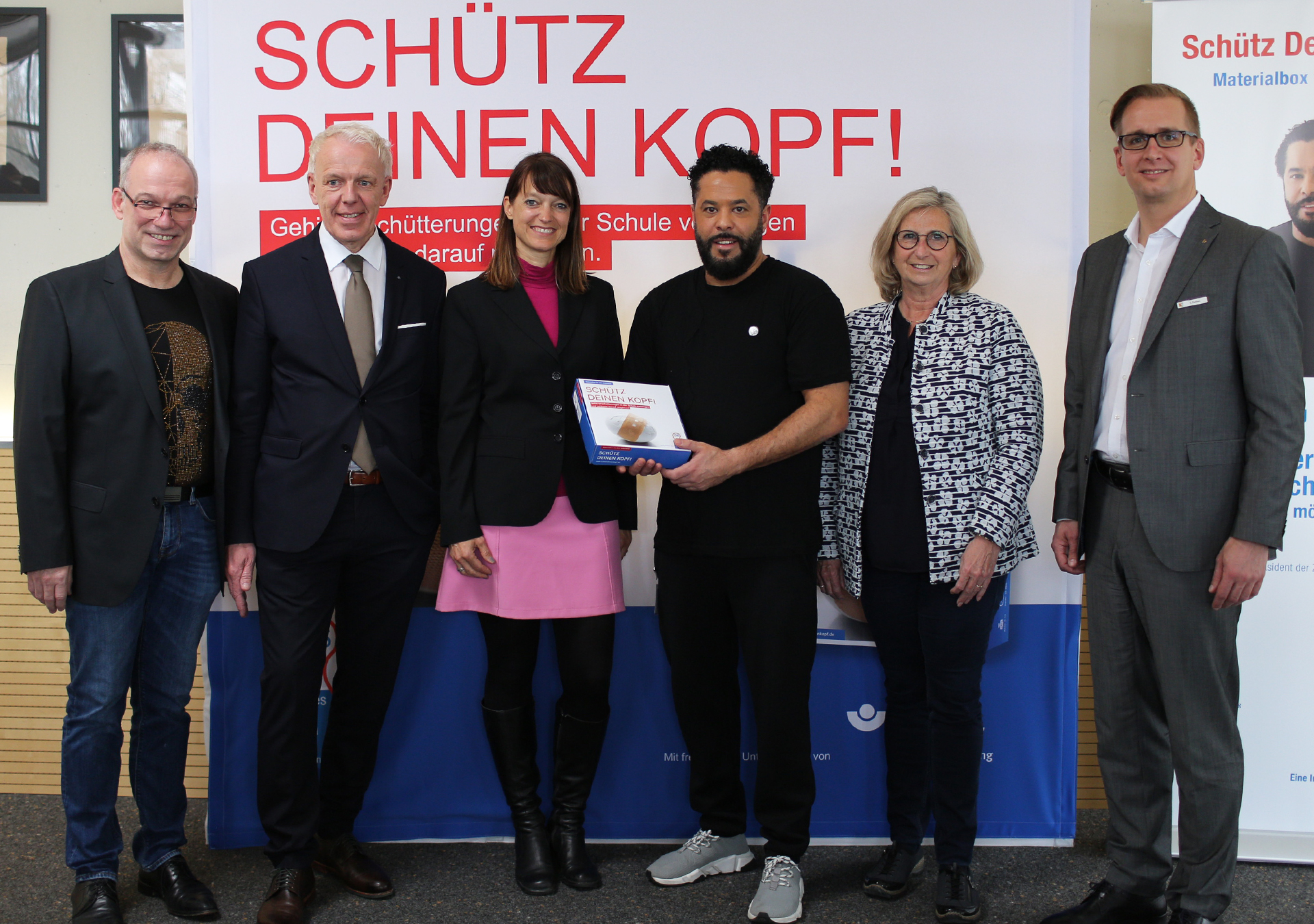 Foto (ZNS): Über den gelungenen Pilotauftakt in Oldenburg freuen sich die Projektbeteiligten die Projektbeteiligten stehen vor dem Banner Schütz Deinen Kopf - Gehirnerschütterungen in der Schule vermeiden, mittig mit der Box steht Adel Tawil als Präsident der ZNS